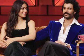 Rashmika Mandanna and Vijay Deverakonda seated on a sofa on a red-lit talk show set, with a pink coffee cup on a side table