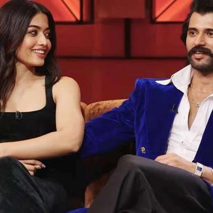 Rashmika Mandanna and Vijay Deverakonda seated on a sofa on a red-lit talk show set, with a pink coffee cup on a side table