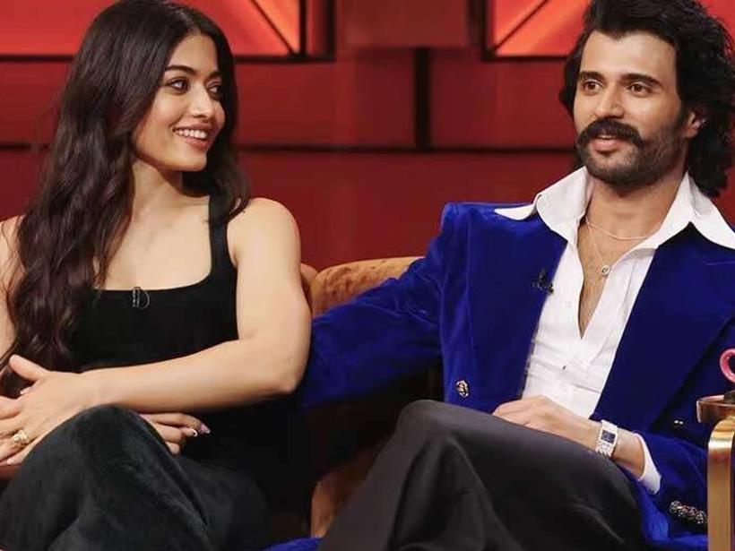 Rashmika Mandanna and Vijay Deverakonda seated on a sofa on a red-lit talk show set, with a pink coffee cup on a side table