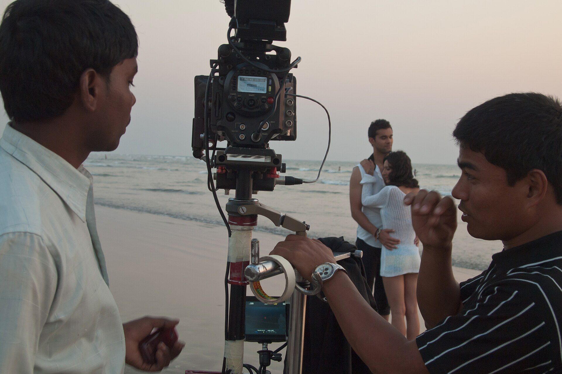 A film camera on a tripod at the water's edge on a beach, with two crew members and a couple standing behind it