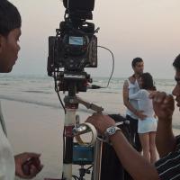 A film camera on a tripod at the water's edge on a beach, with two crew members and a couple standing behind it