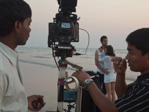 A film camera on a tripod at the water's edge on a beach, with two crew members and a couple standing behind it