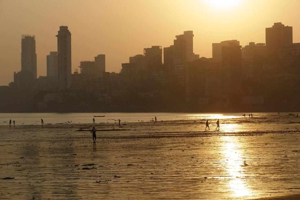 Silhouetted people walk on a wet beach at sunset, with Mumbai's high-rise skyline behind them.