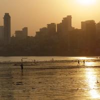 Silhouetted people walk on a wet beach at sunset, with Mumbai's high-rise skyline behind them.