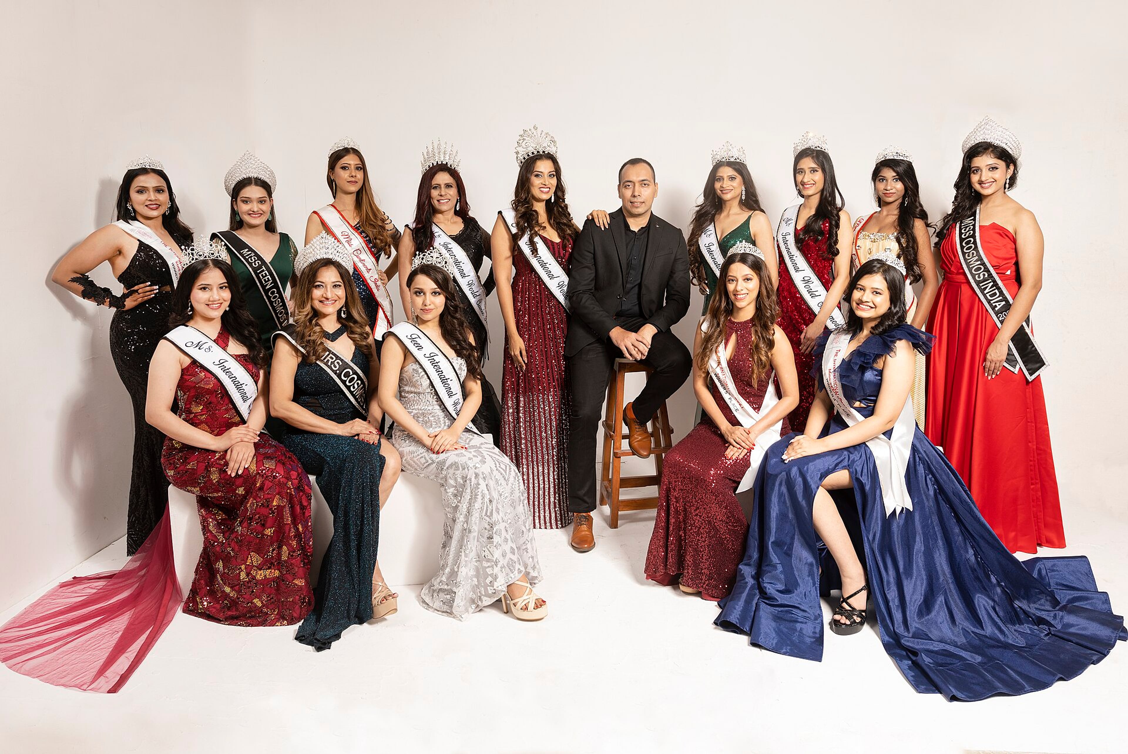 Group of women in gowns and sashes posing with a man in a dark suit against a white studio backdrop