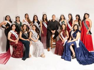 Group of women in gowns and sashes posing with a man in a dark suit against a white studio backdrop