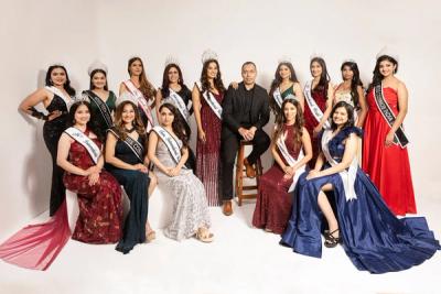 Group of women in gowns and sashes posing with a man in a dark suit against a white studio backdrop