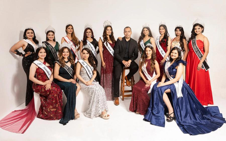 Group of women in gowns and sashes posing with a man in a dark suit against a white studio backdrop