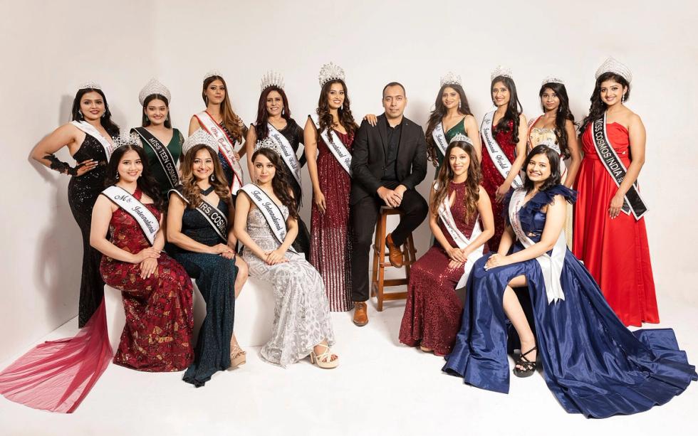 Group of women in gowns and sashes posing with a man in a dark suit against a white studio backdrop