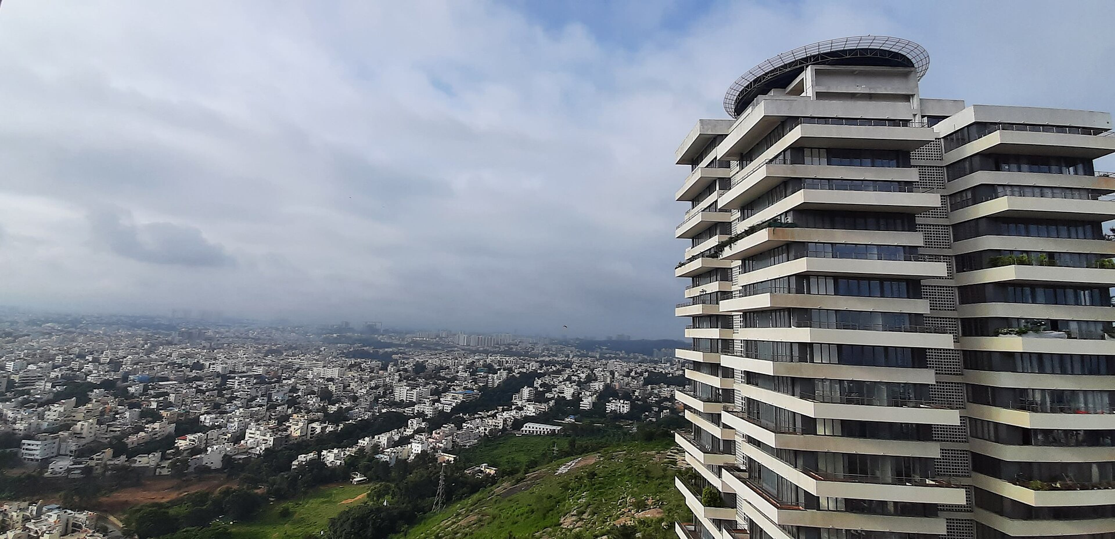 Panoramic view over a dense city of low buildings, with a tall balconied tower on a green hillside under a cloudy sky