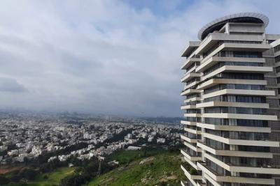 Panoramic view over a dense city of low buildings, with a tall balconied tower on a green hillside under a cloudy sky