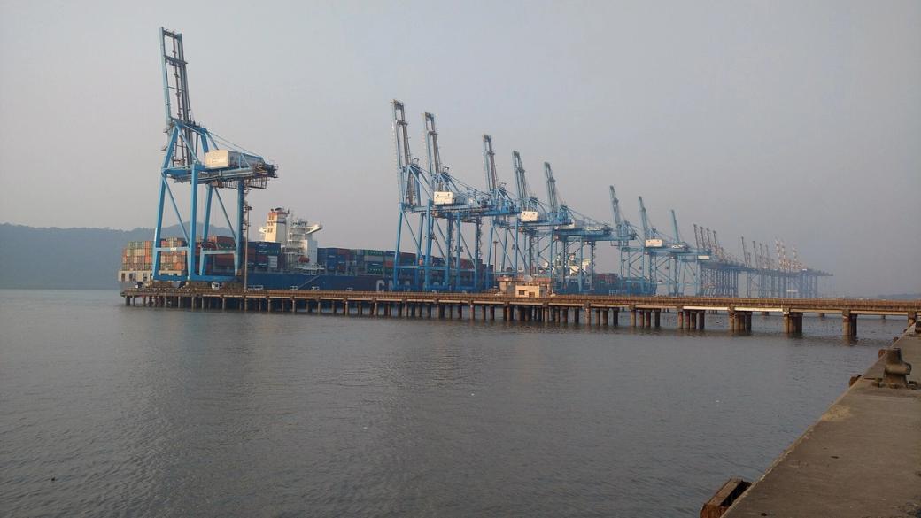 Blue gantry cranes unload shipping containers from a cargo ship at a port beside calm, hazy water.