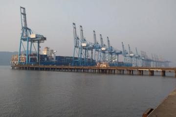 Blue gantry cranes unload shipping containers from a cargo ship at a port beside calm, hazy water.