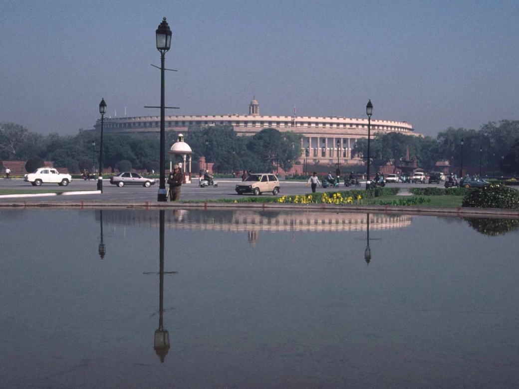 Parliament building in New Delhi seen across a reflecting pool, with cars and pedestrians on the road beside it