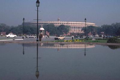 Parliament building in New Delhi seen across a reflecting pool, with cars and pedestrians on the road beside it