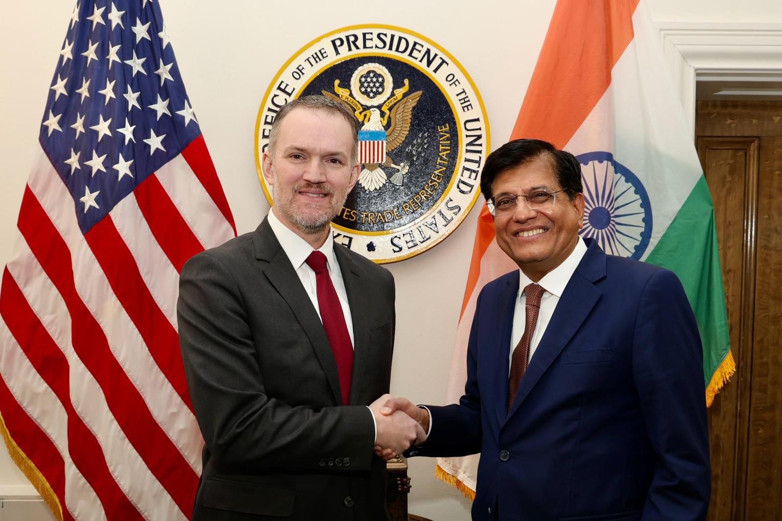 Two men in suits shake hands in front of a US flag, an Indian flag and an Office of the US Trade Representative seal