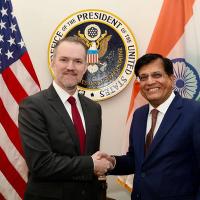 Two men in suits shake hands in front of a US flag, an Indian flag and an Office of the US Trade Representative seal