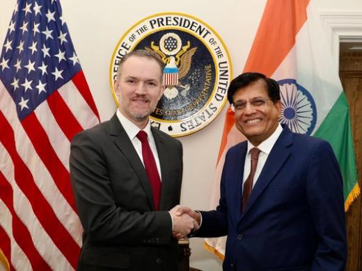 Two men in suits shake hands in front of a US flag, an Indian flag and an Office of the US Trade Representative seal