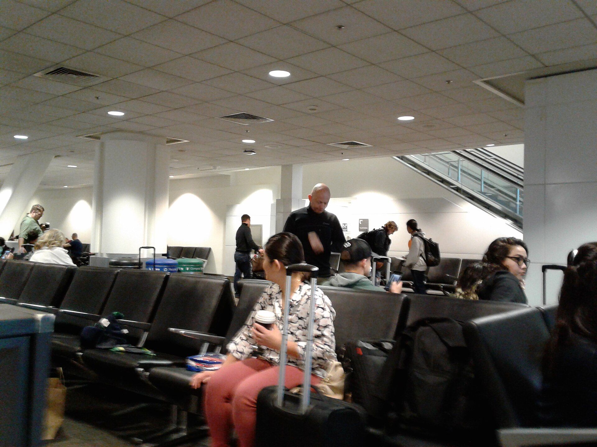 Travellers sit and stand in an airport waiting area with black seats and overhead ceiling lights.