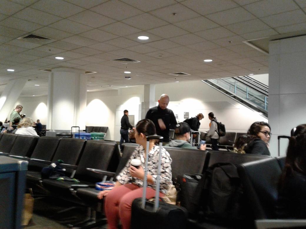 Travellers sit and stand in an airport waiting area with black seats and overhead ceiling lights.