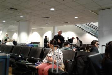 Travellers sit and stand in an airport waiting area with black seats and overhead ceiling lights.