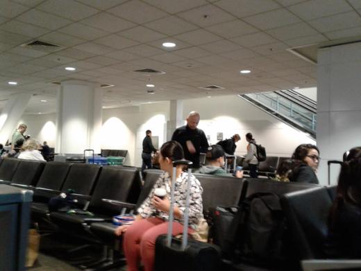 Travellers sit and stand in an airport waiting area with black seats and overhead ceiling lights.