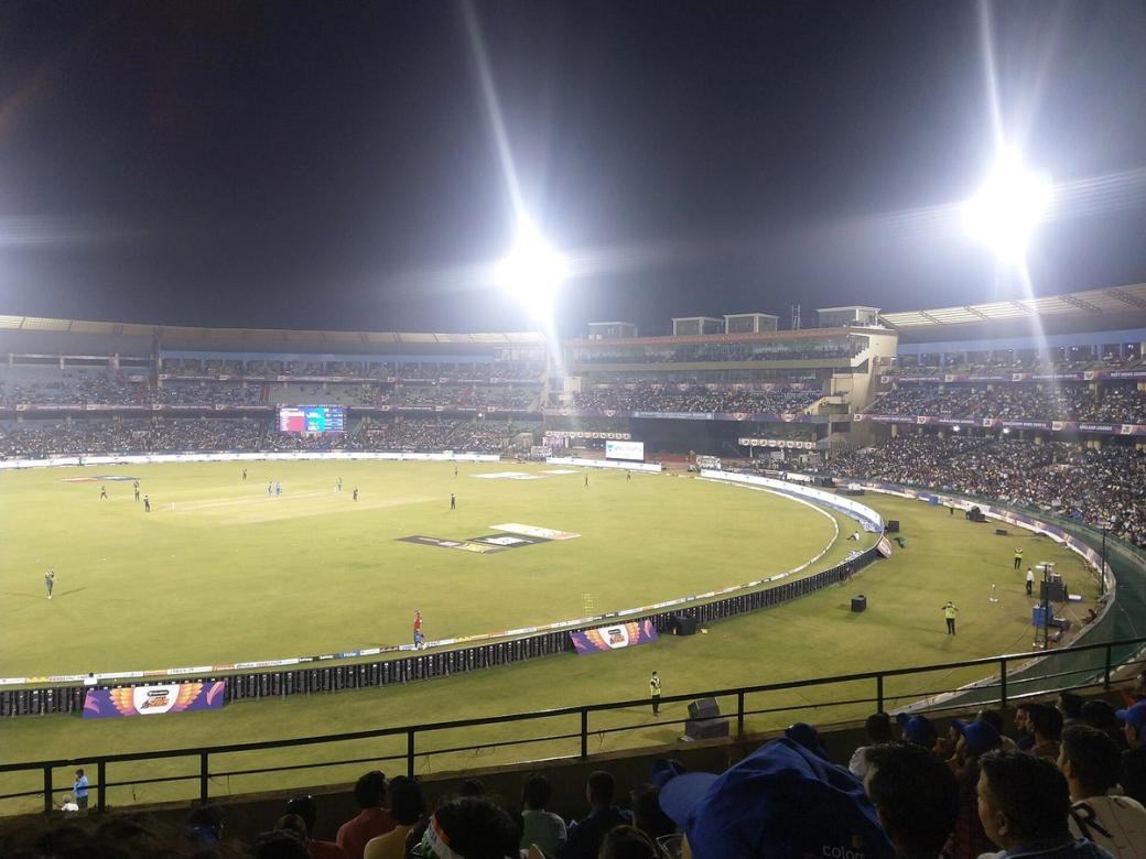 Floodlit night cricket match in progress, with players on the field and spectators filling the stands.
