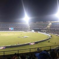 Floodlit night cricket match in progress, with players on the field and spectators filling the stands.