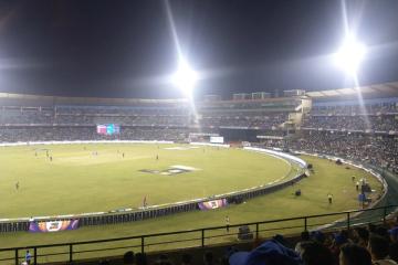 Floodlit night cricket match in progress, with players on the field and spectators filling the stands.