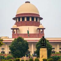 Central Wing of the Supreme Court of India with its domed tower, a statue on a plinth and people at the entrance.