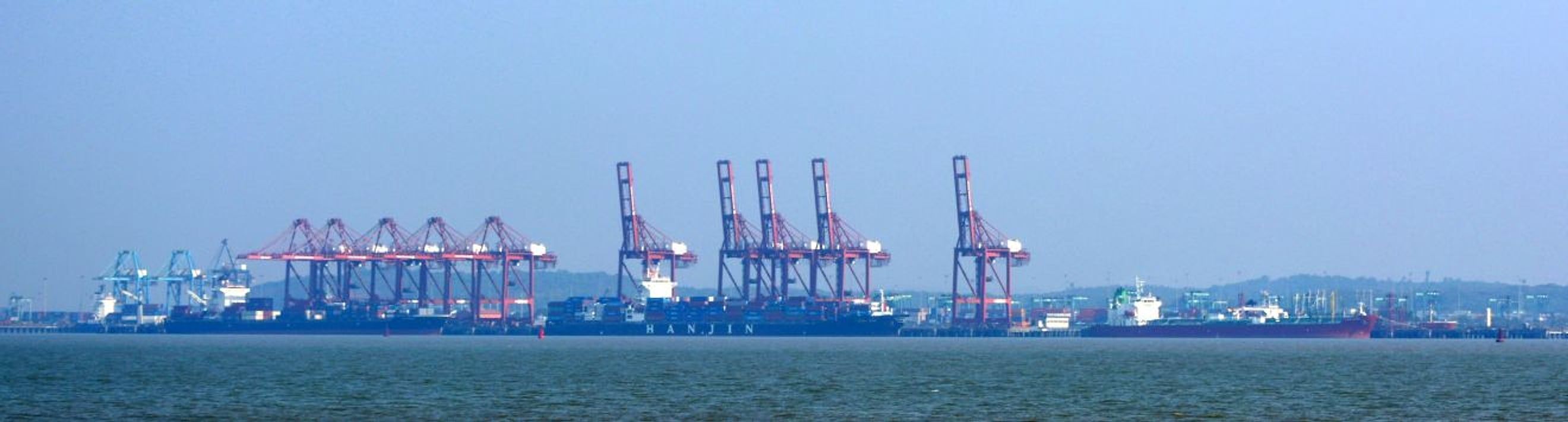 Row of red container cranes and cargo ships docked at a port, seen across the water under a hazy blue sky.