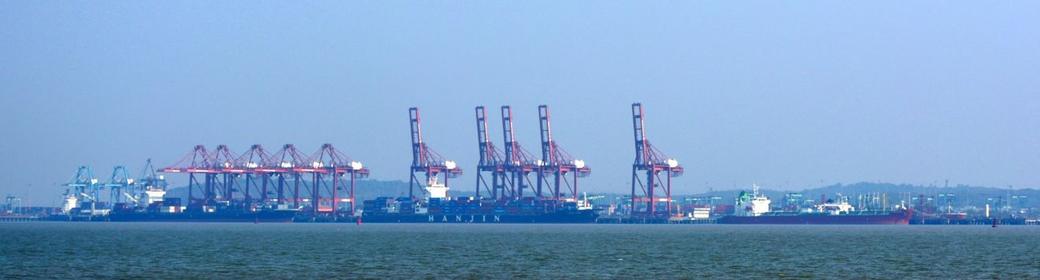 Row of red container cranes and cargo ships docked at a port, seen across the water under a hazy blue sky.