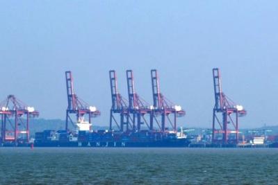 Row of red container cranes and cargo ships docked at a port, seen across the water under a hazy blue sky.
