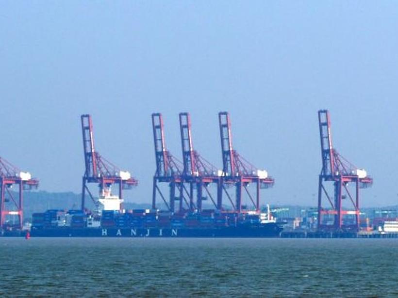 Row of red container cranes and cargo ships docked at a port, seen across the water under a hazy blue sky.