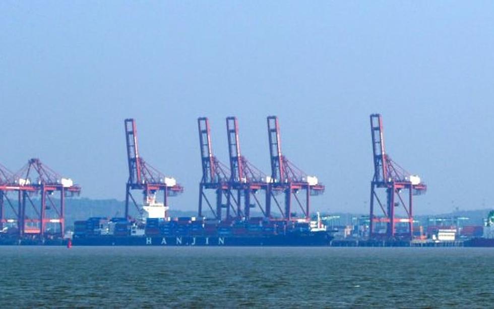 Row of red container cranes and cargo ships docked at a port, seen across the water under a hazy blue sky.