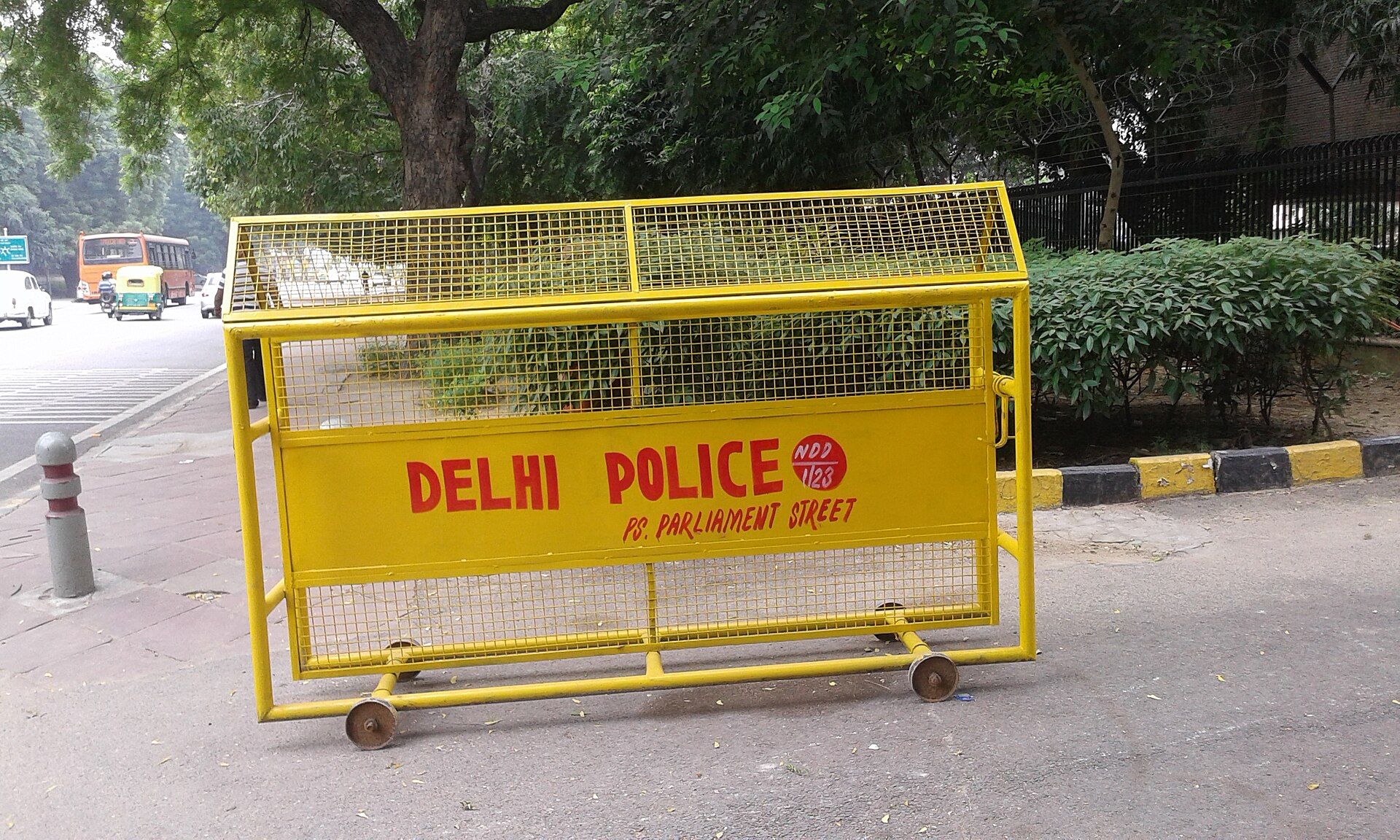 Yellow Delhi Police barricade on a pavement beside a road, with trees, a hedge and a bus in the background