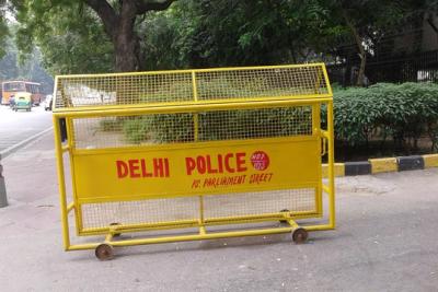 Yellow Delhi Police barricade on a pavement beside a road, with trees, a hedge and a bus in the background