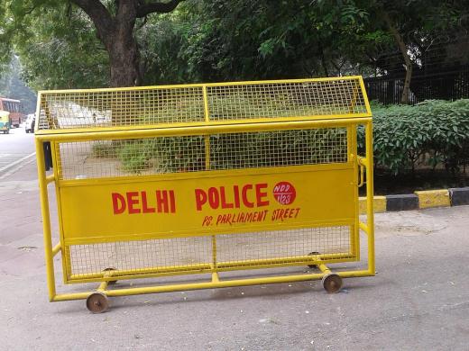 Yellow Delhi Police barricade on a pavement beside a road, with trees, a hedge and a bus in the background