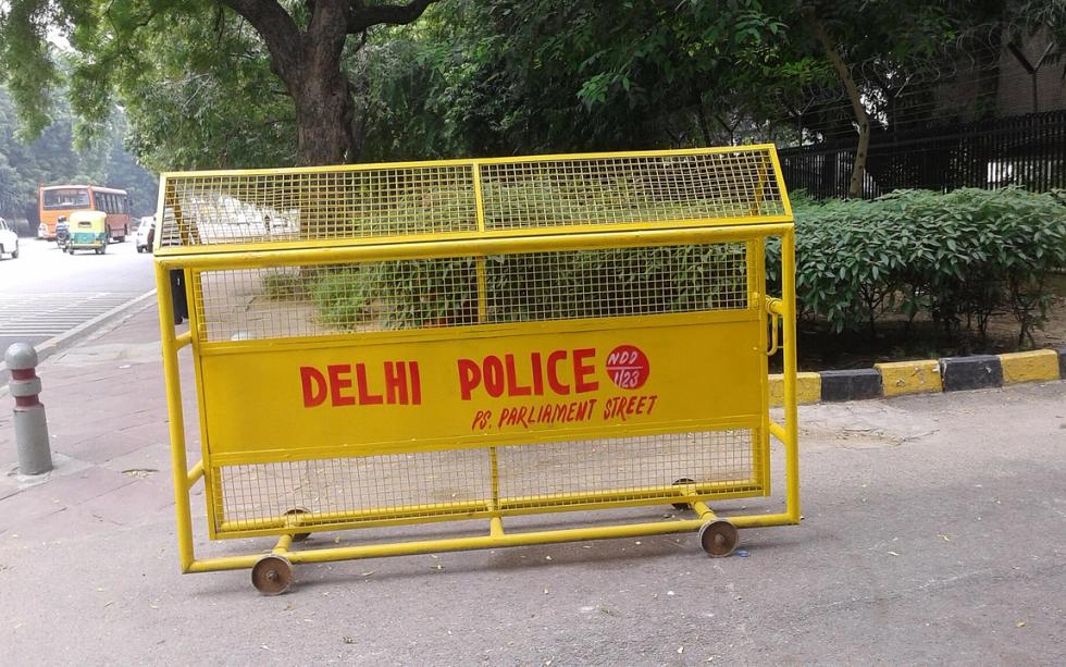 Yellow Delhi Police barricade on a pavement beside a road, with trees, a hedge and a bus in the background