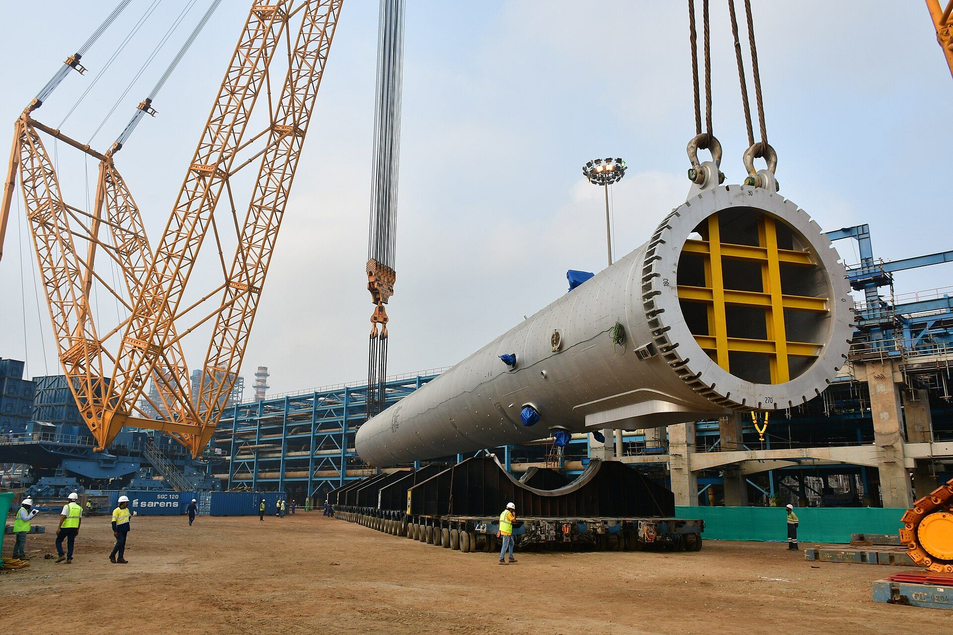 Workers in hard hats beside a huge steel reactor vessel slung from cranes at a refinery construction site