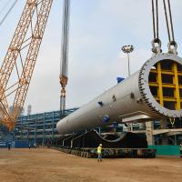 Workers in hard hats beside a huge steel reactor vessel slung from cranes at a refinery construction site