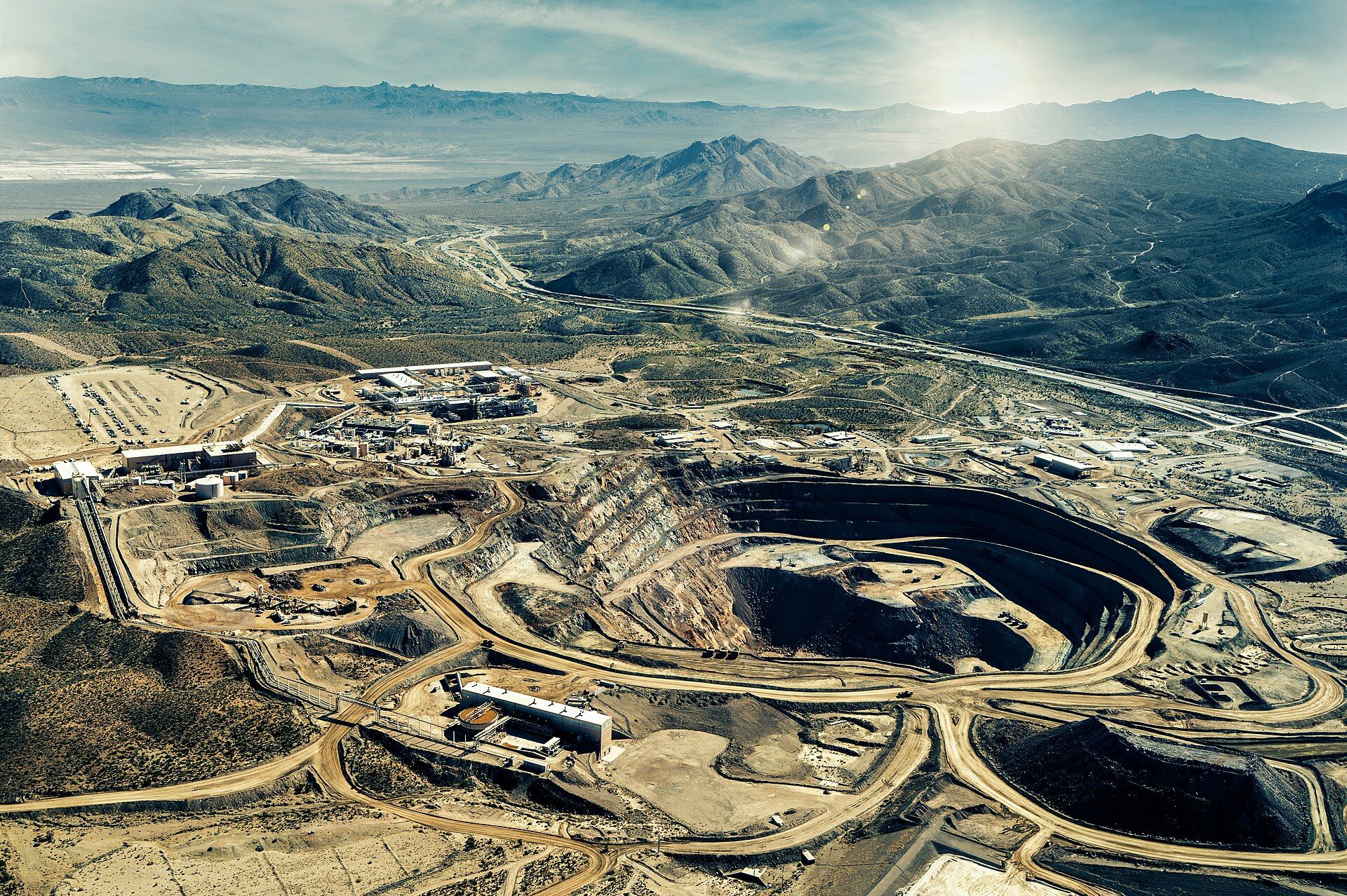 Aerial view of an open-pit mine and processing buildings in a desert landscape with mountains and a highway.