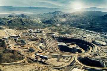 Aerial view of an open-pit mine and processing buildings in a desert landscape with mountains and a highway.