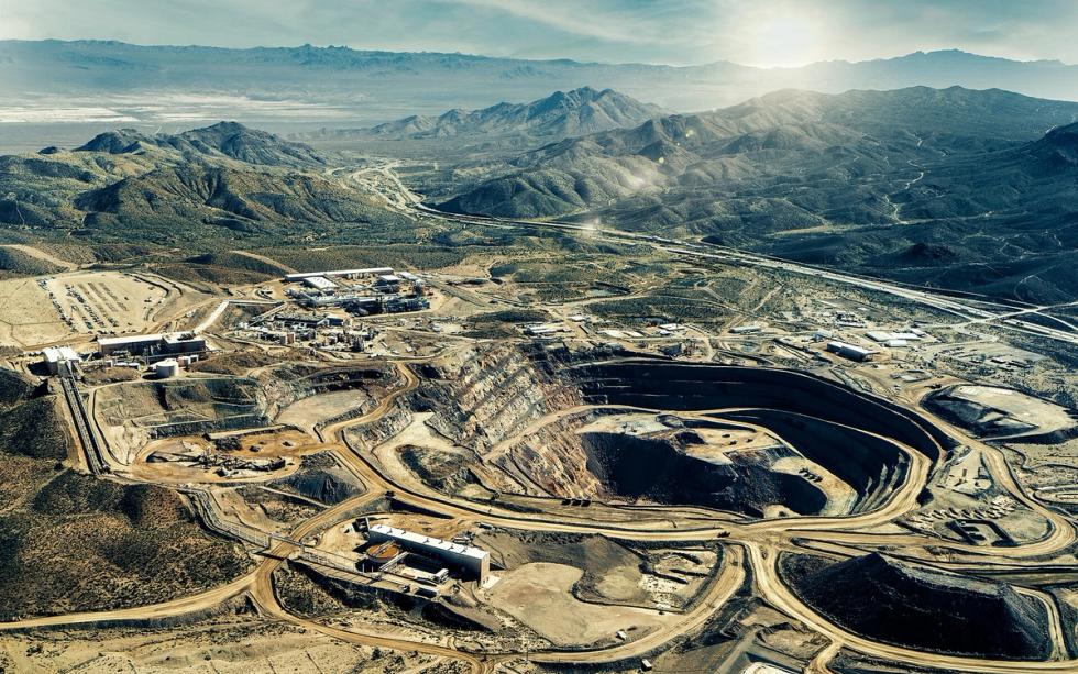 Aerial view of an open-pit mine and processing buildings in a desert landscape with mountains and a highway.