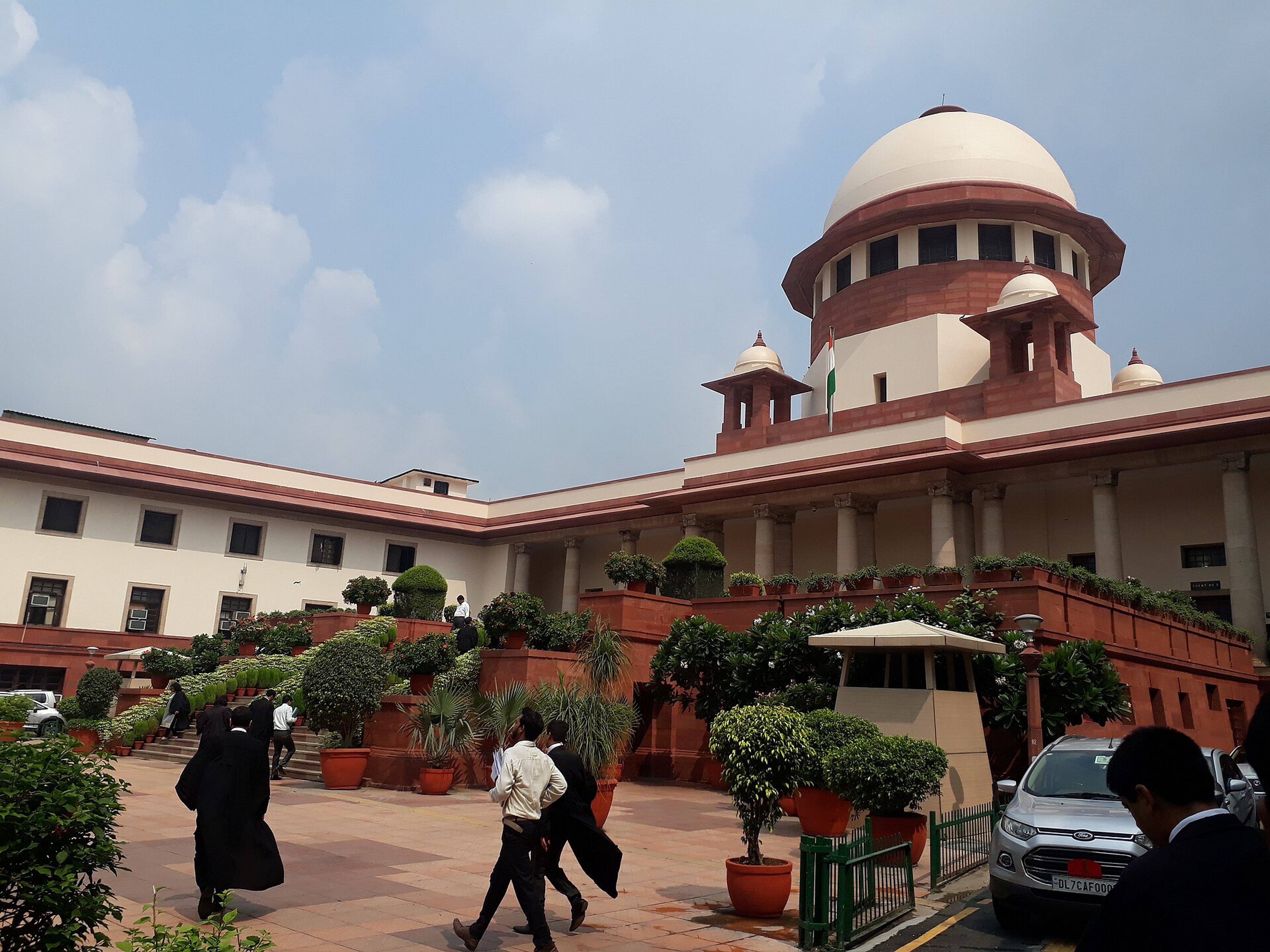 People walk across a paved area before the Supreme Court of India building, with potted plants and parked cars.
