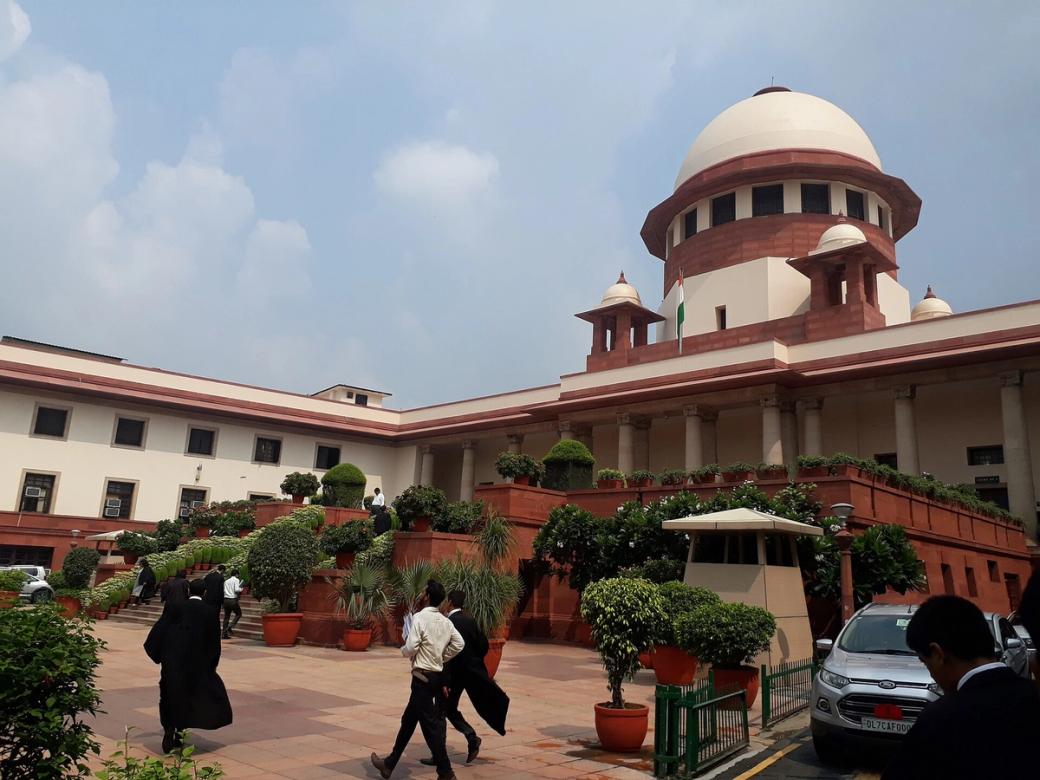 People walk across a paved area before the Supreme Court of India building, with potted plants and parked cars.