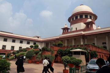People walk across a paved area before the Supreme Court of India building, with potted plants and parked cars.