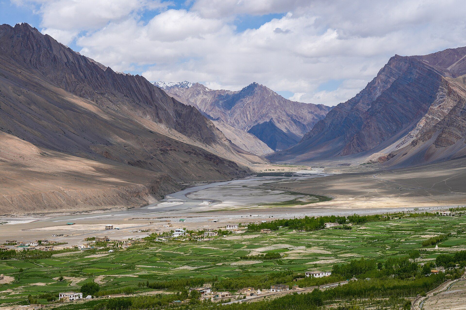 Green fields of a valley village with a winding river and snow-streaked mountains under a cloudy sky.