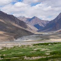 Green fields of a valley village with a winding river and snow-streaked mountains under a cloudy sky.