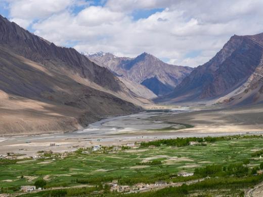 Green fields of a valley village with a winding river and snow-streaked mountains under a cloudy sky.